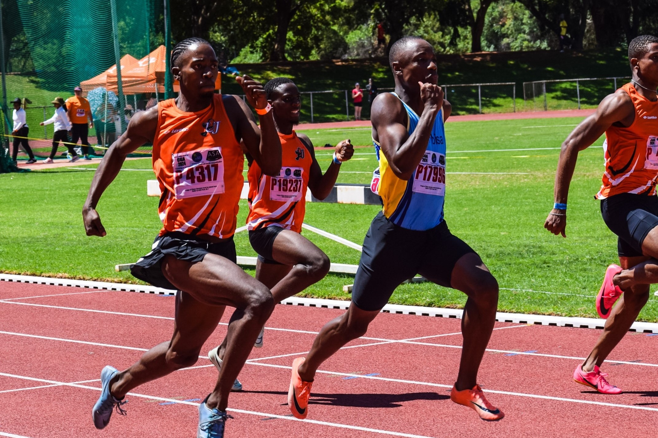 Four boys running, and three of them are wearing orange (top) and black (shorts) representing UJ Athletics team and the other one is in blue top and black shorts