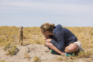 Person crouching on sandy, dry ground in a barren field, facing a standing meerkat nearby.
