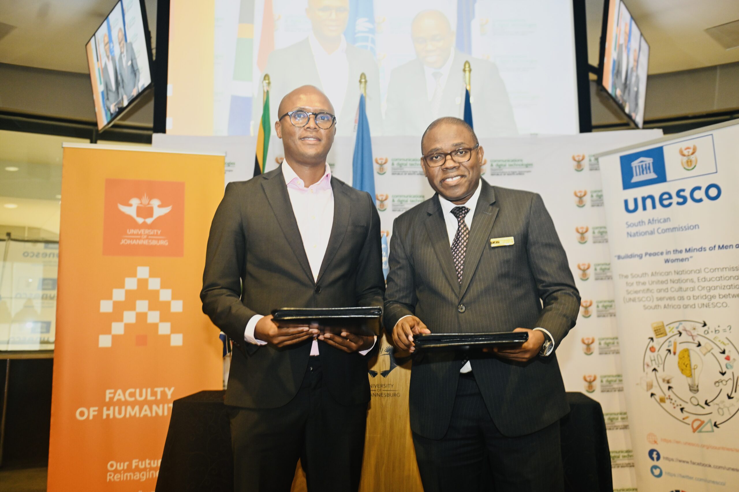Two men in suits posing and holding tablets at a formal event, with UNESCO and university banners in the background.