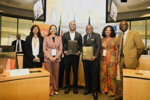 Six professionals posing for a group photo at a signing ceremony, two men in suits holding black folders and four colleagues smiling in a conference room behind banners and flags to mark the event, all wearing conference lanyards and formal attire.