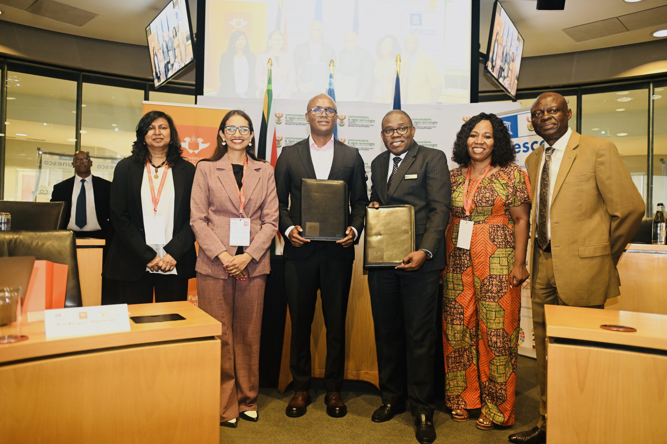 Six professionals posing for a group photo at a signing ceremony, two men in suits holding black folders and four colleagues smiling in a conference room behind banners and flags to mark the event, all wearing conference lanyards and formal attire.