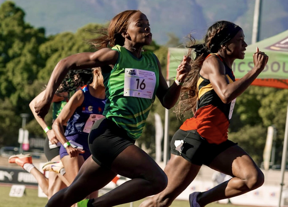 Group of female sprinters racing on a track, wearing colorful uniforms and bib numbers, mid-stride with a green-clad lead runner.