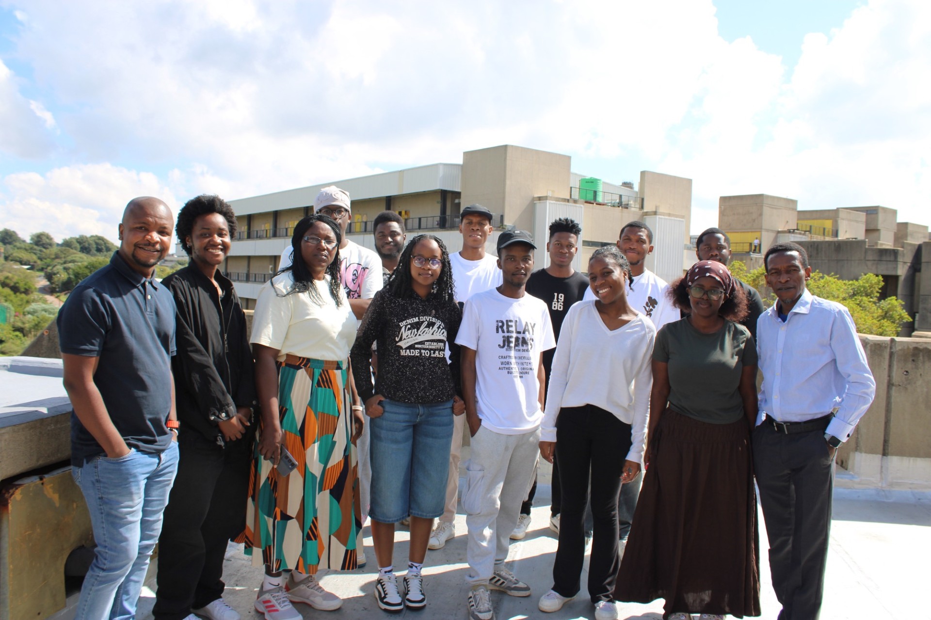 Group of smiling people standing together on a rooftop, posing for a photo outdoors on a sunny day.