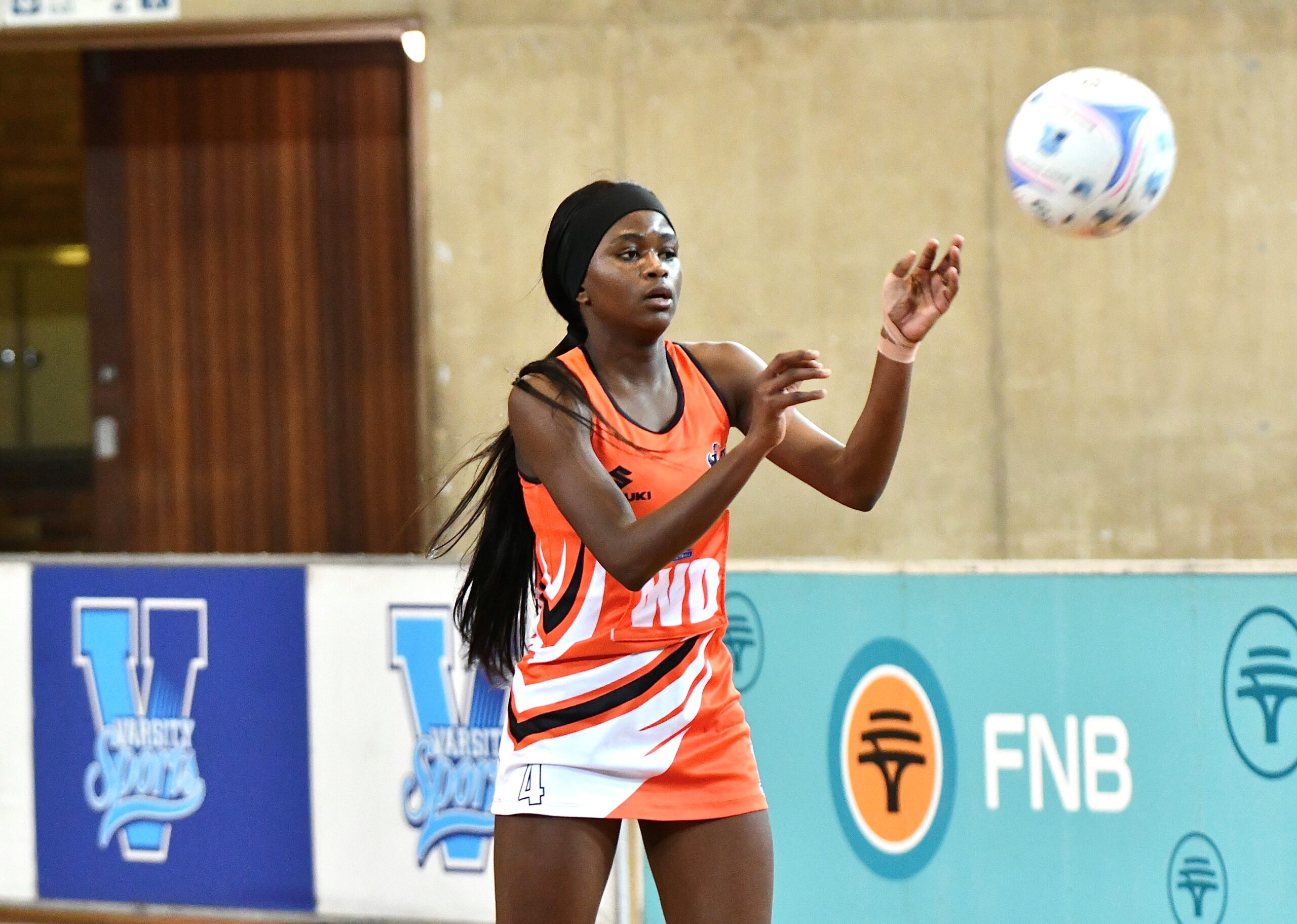 Female volleyball player in an orange uniform reaching for a ball indoors, wearing a black headband and long hair.