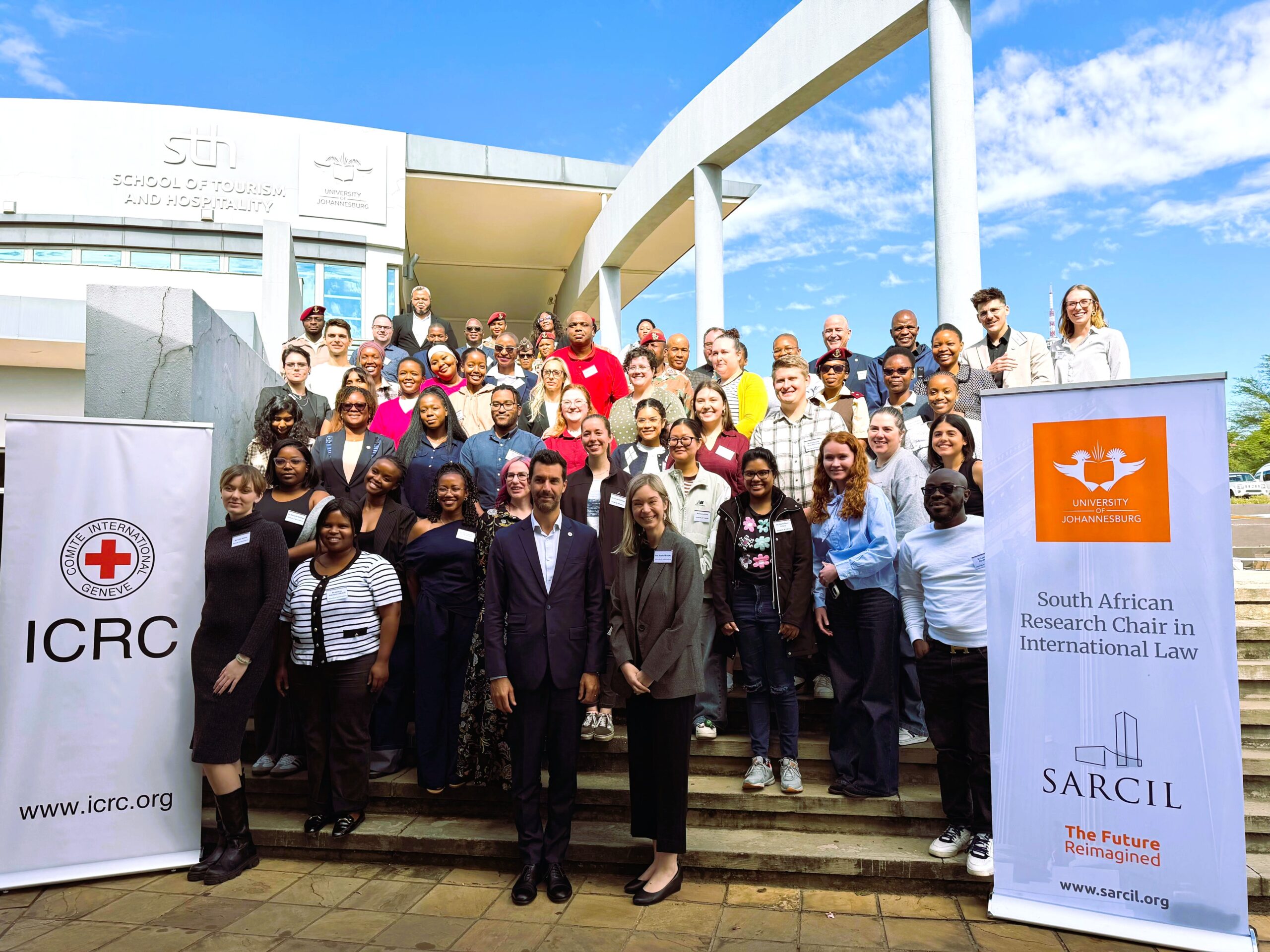 Large diverse group posing on steps outside a university building, with banners for ICRC and the South African Research Chair in International Law visible at sides.