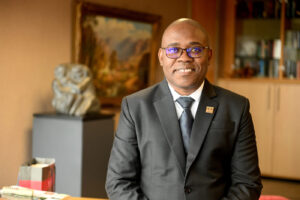 Smiling man in a dark suit and glasses seated in a warmly lit office, with bookshelves in the background.