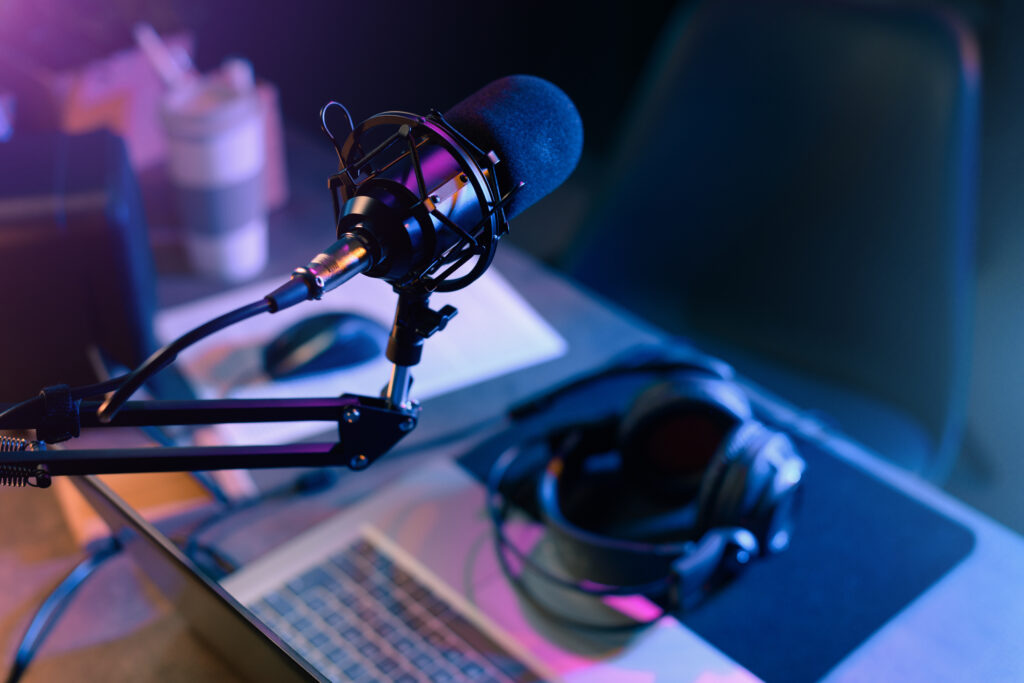 Professional studio microphone mounted on a boom arm in a color-lit recording setup with a laptop nearby and headphones on the desk.