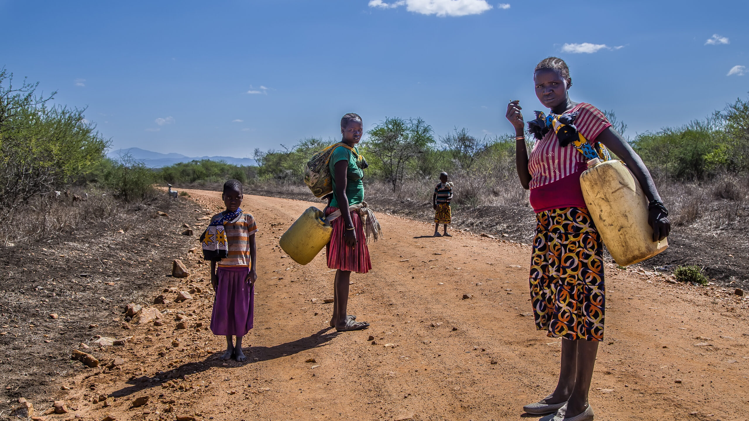 People stand on a dusty rural road with plastic jerry cans and a backpack in a dry, scrubby landscape under a blue sky .