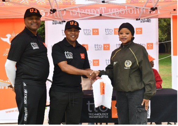 Three people under an orange canopy at an outdoor event; two men in black shirts shake hands with a woman in a green jacket in front of a logoed backdrop.