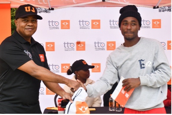 Two men shake hands at a community event under an orange canopy; the man on the left wears a black polo and a U cap, the man on the right wears a gray sweatshirt and a black beanie, holding orange materials and a bag.
