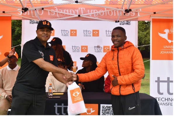 Two men shake hands under an orange event tent, exchanging a white bag with an orange logo; one wears a black shirt and cap, the other an orange puffer jacket.