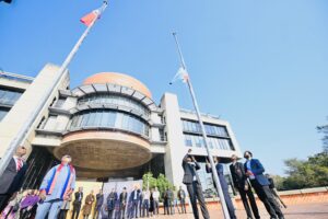 Group of professionals in suits standing outside a modern building with flagpoles and an orange-brick facade near the entrance.