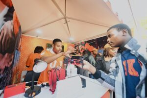 Young man in an orange shirt demonstrates a red device to a visitor at a tech booth under a white canopy.
