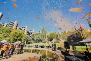 Outdoor celebration with orange confetti raining over a stage and audience beneath a blue sky, with colorful flags and buildings in the background.