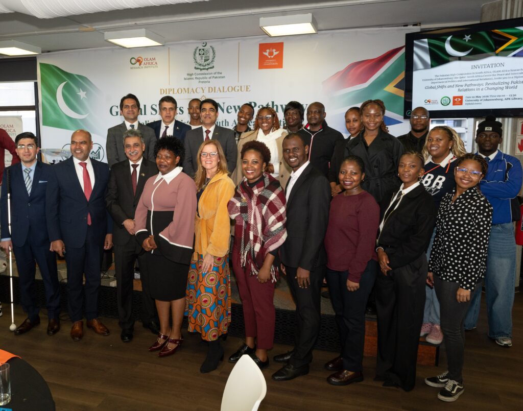 Diverse group posing for a group photo at a diplomacy dialogue event, with banners and flags in the background.