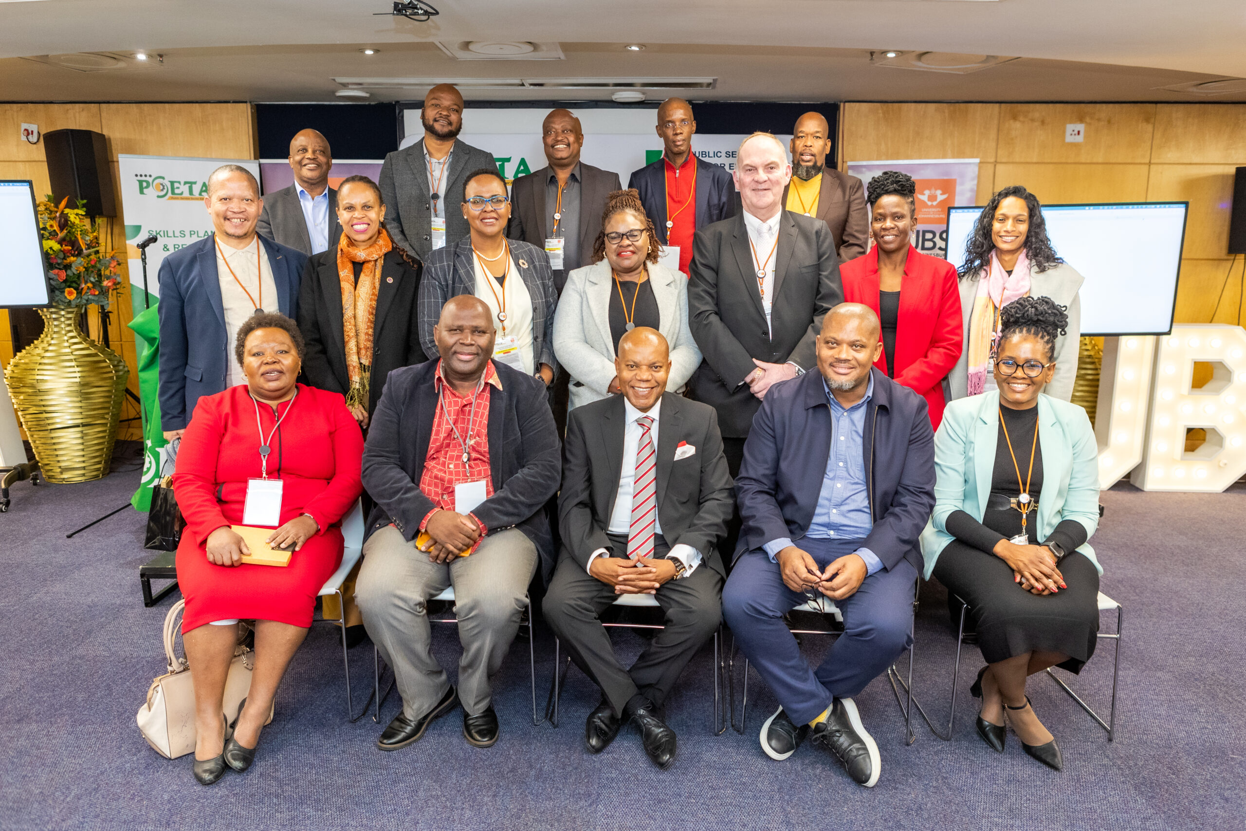 Group of professionally dressed people posing for a group photo at a conference, with name badges and banners in the background.