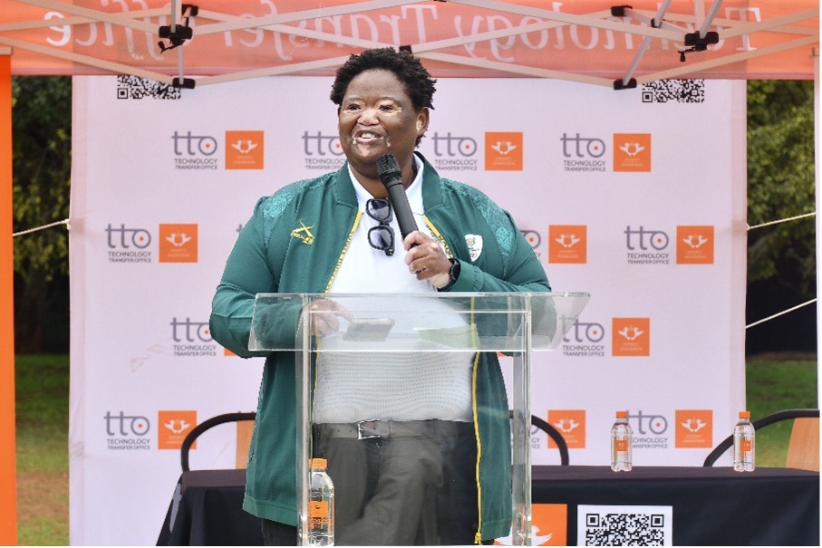 Woman in a green jacket speaks into a microphone at a clear podium during an outdoor tech event, with a branded backdrop and water bottles on the table.