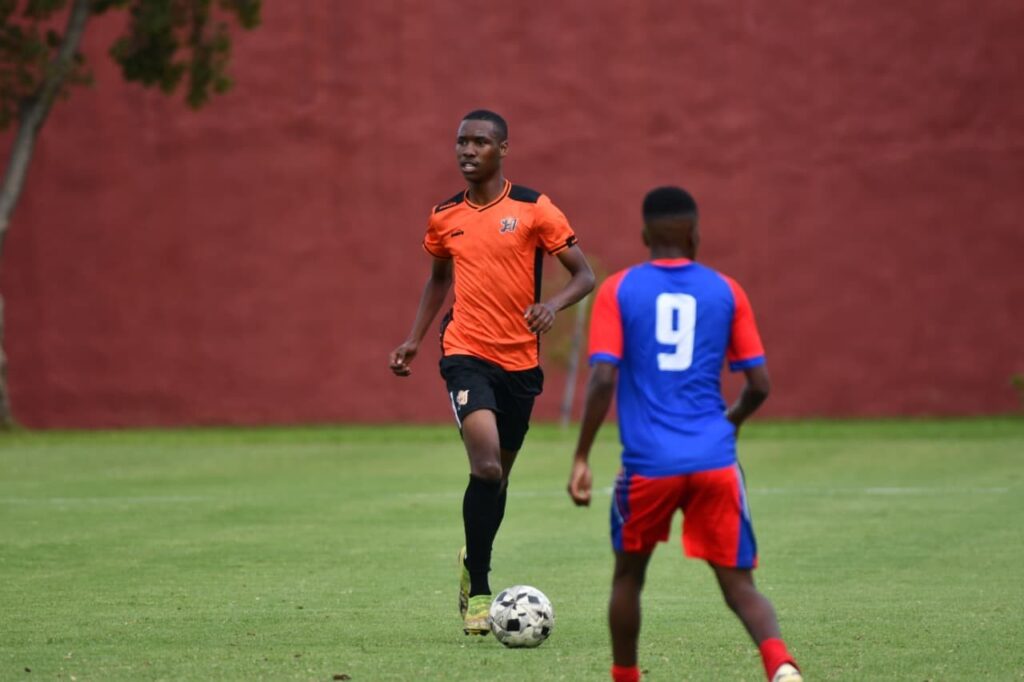 Soccer players on a green field; an orange-jersey player dribbles the ball while a blue-and-red defender approaches.