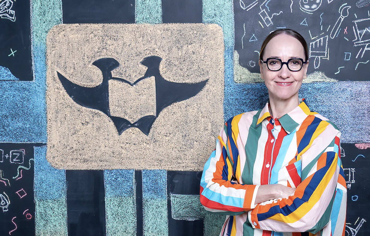 Smiling woman with glasses in a multicolored striped shirt, arms crossed, standing beside a chalkboard mural with a beige slab featuring a black bat emblem.