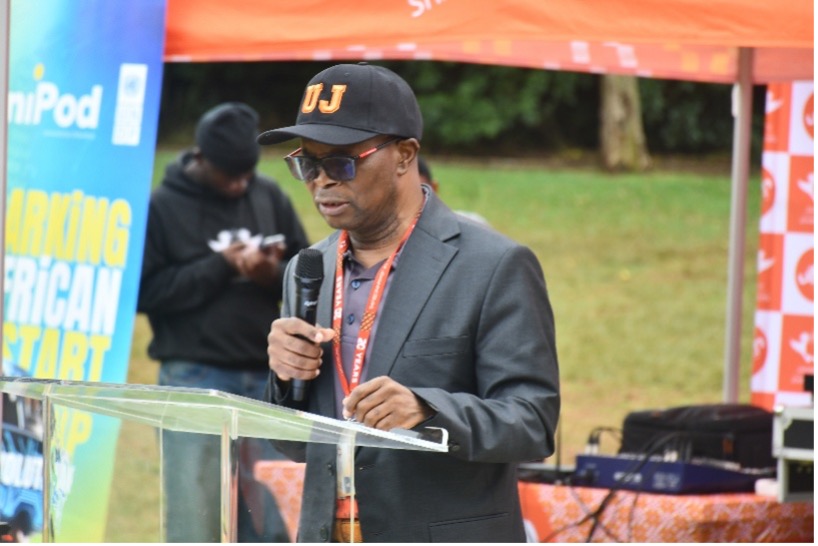 Man in a dark suit and a DJ cap speaks into a microphone at an outdoor podium, with colorful banners behind him.