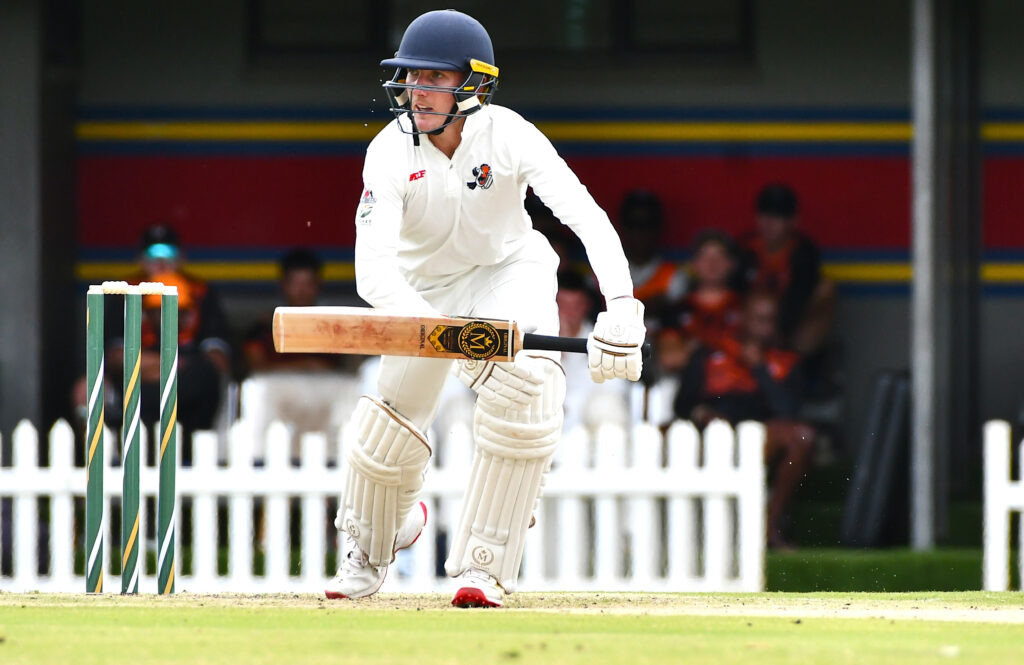 Cricket batsman in white gear drives the ball, helmet on, near the stumps on the left.
