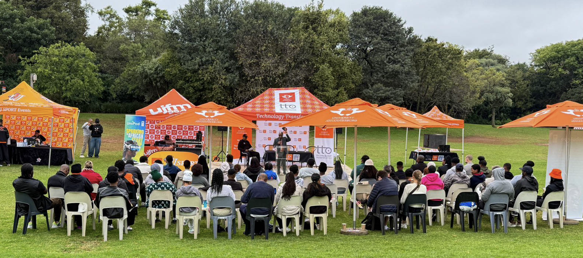 Outdoor community event in a park with bright orange tents, a small stage, and seated audience watching a speaker.