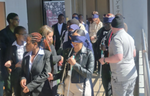 Diverse group of blindfolded participants lined up outside a building, some guiding each other during an activity.