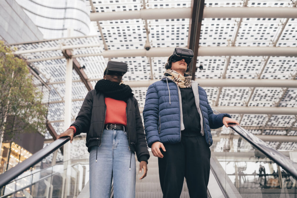 Two people on an escalator wearing VR headsets in a glass-walled mall.