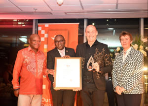 Four people at an awards ceremony pose with a framed certificate and a glass trophy against an orange backdrop.