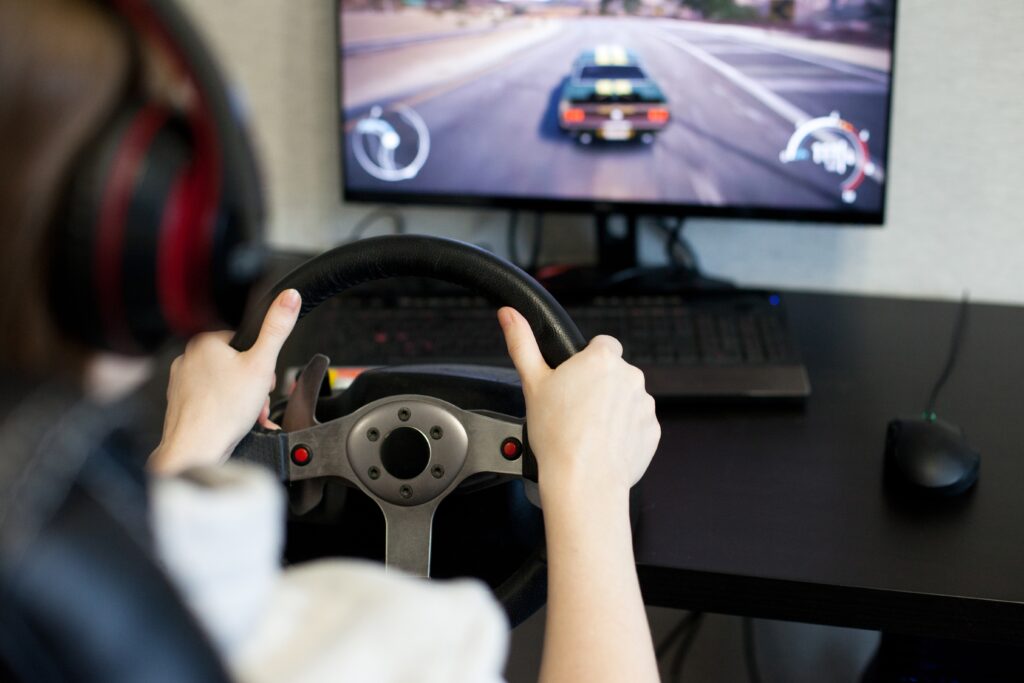 Person grips a car racing wheel controller while playing a driving video game, with a blurred racing game on the screen in the background.