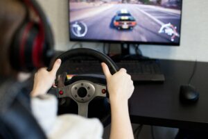 Person grips a car racing wheel controller while playing a driving video game, with a blurred racing game on the screen in the background.