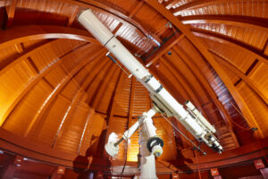 Interior of an observatory dome with orange wooden ribs and a large white telescope mounted on a central support,