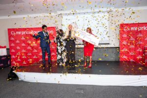 Group of four people celebrating on stage as gold confetti falls; one person holds a large ceremonial check for 1,000,000 rand with a Mr Price Foundation backdrop.