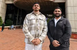 Two men stand outdoors in front of a modern building with a brick-paved plaza: one in a beige plaid shirt, the other in a black zip-up jacket, both smiling.