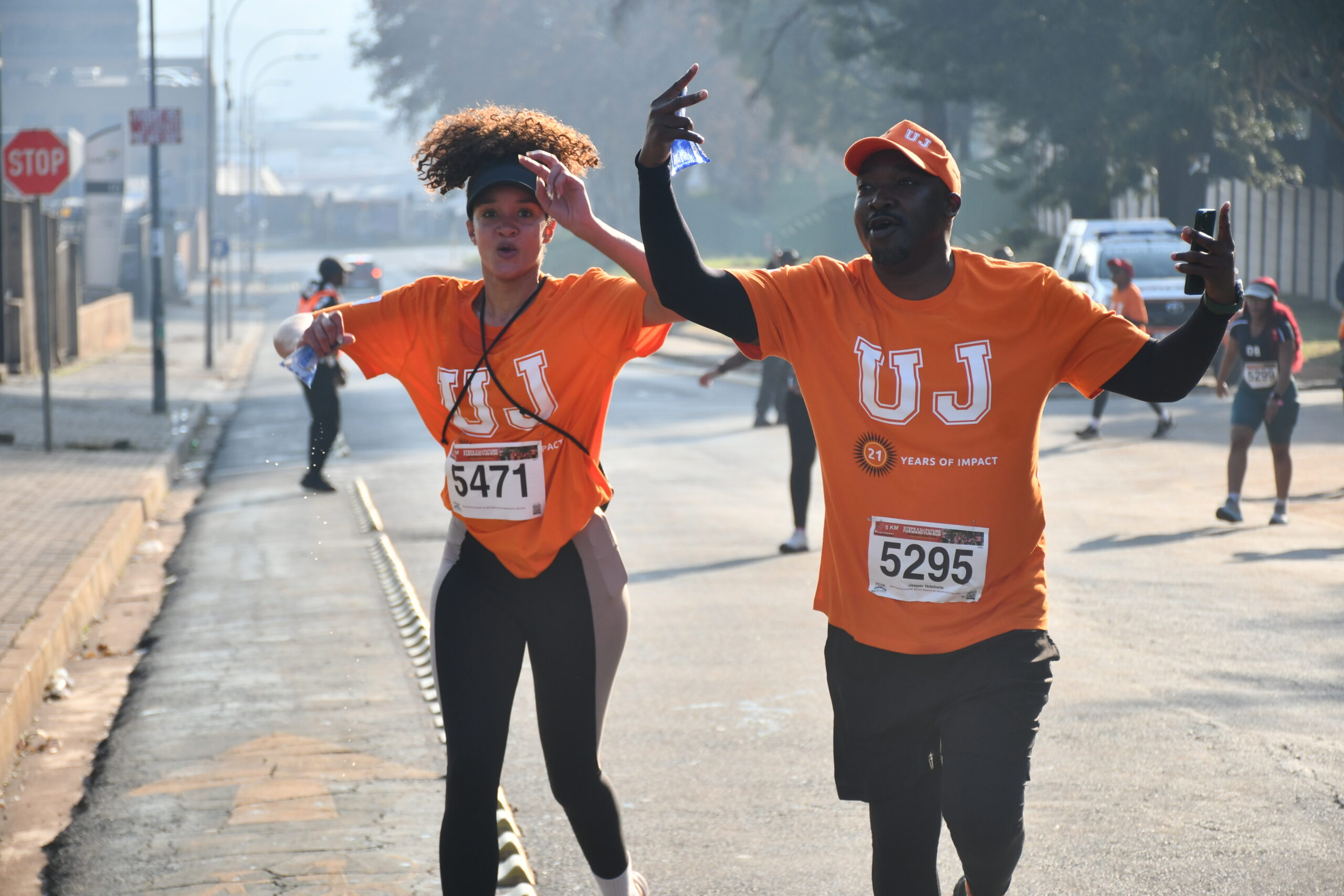 Two runners in orange shirts with race bibs 5471 and 5295 cheer and run toward the camera on a city street, raising their arms in celebration.