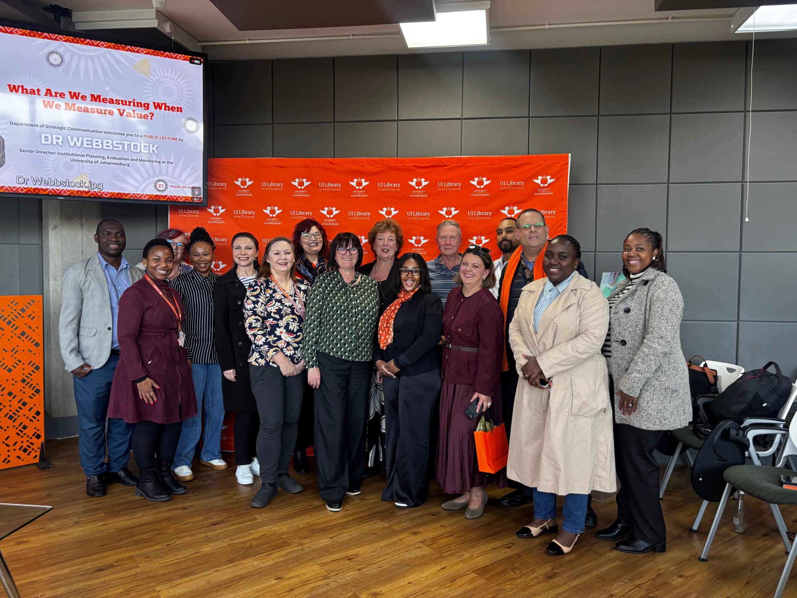 Group of diverse adults posing for a photo in front of an orange UJ Library backdrop at a professional event, smiling at the camera.