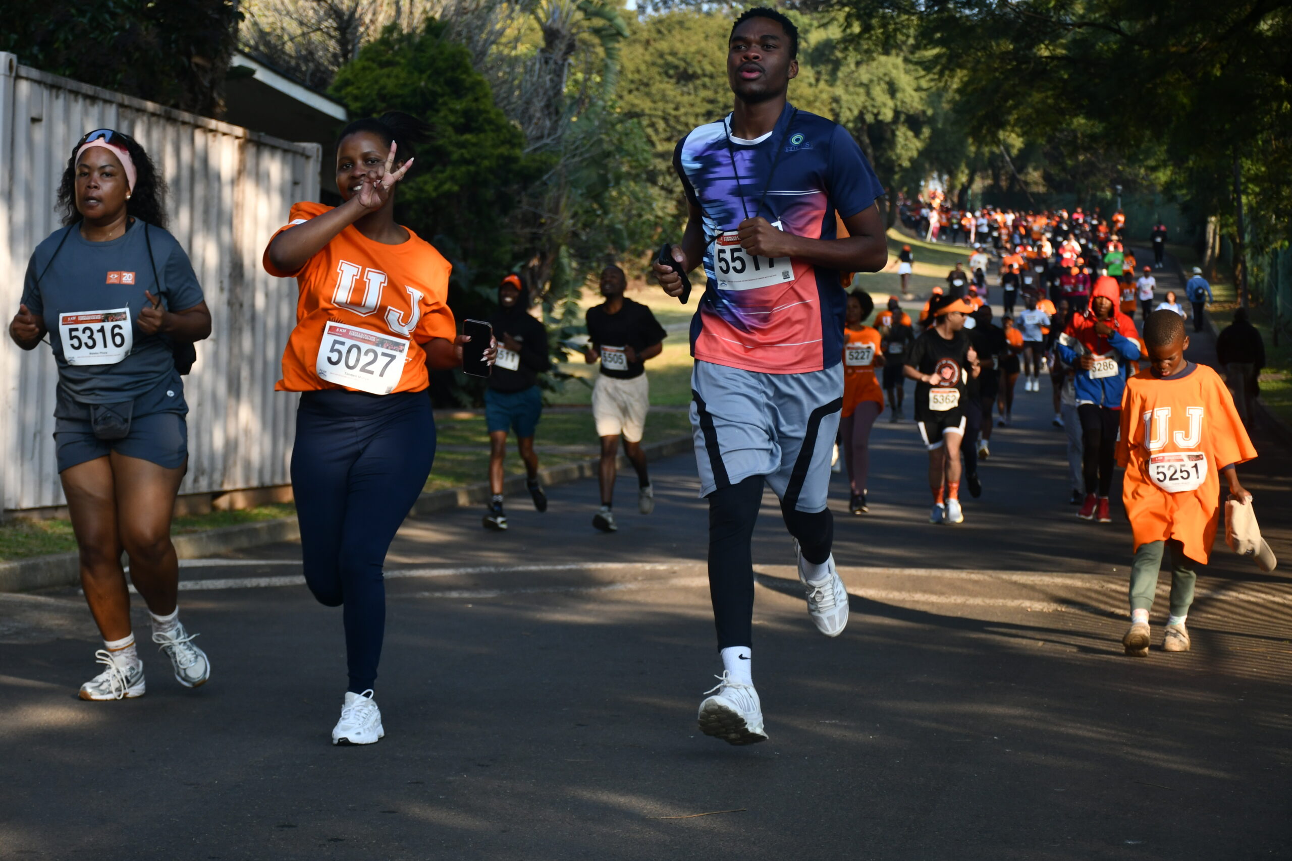 Front runners in a street race: two women and a man in athletic gear, bib numbers visible, running on a tree-lined road with a crowd behind them.