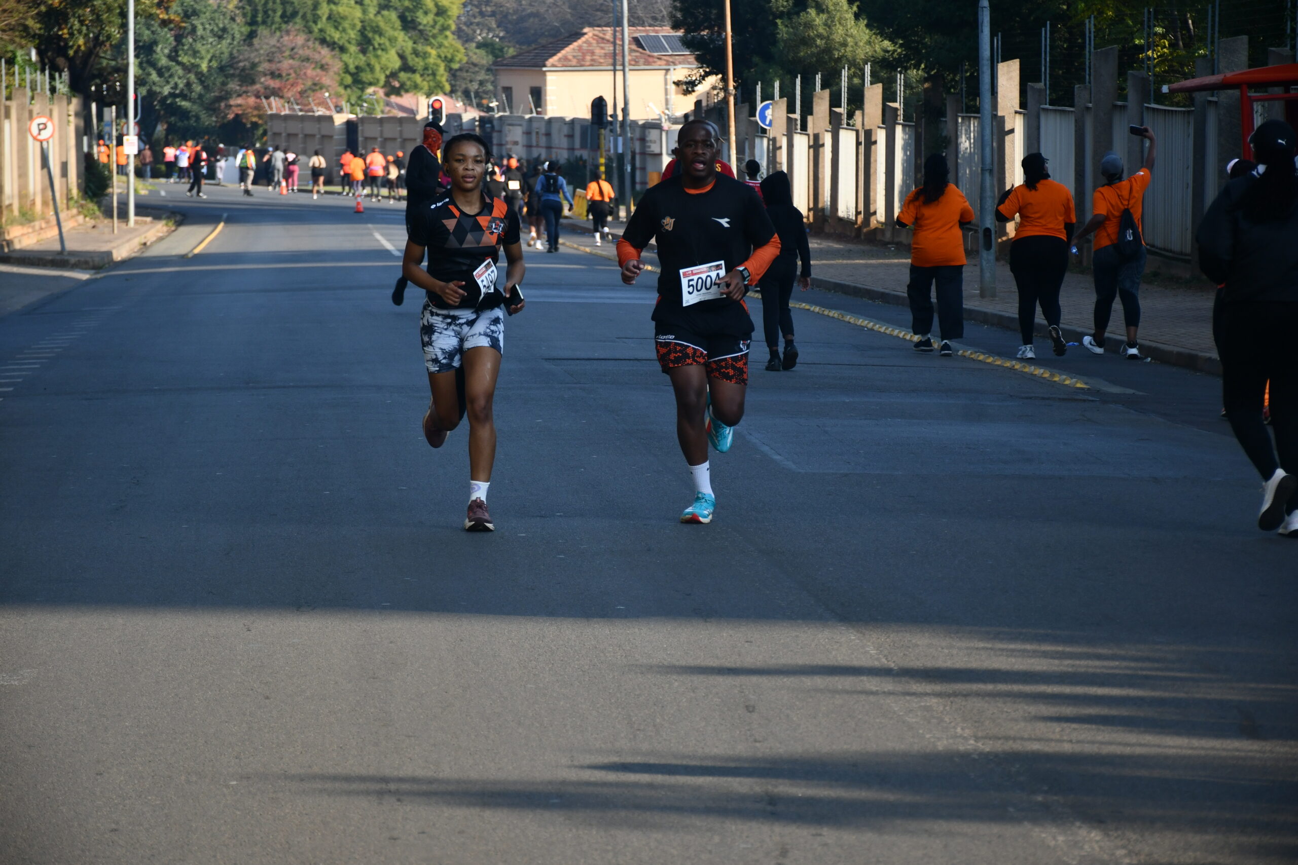 Two runners lead a street race on a city road, with spectators and orange-shirt volunteers lining the route.