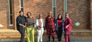 Six adults standing together outside a brick building, smiling for the camera.