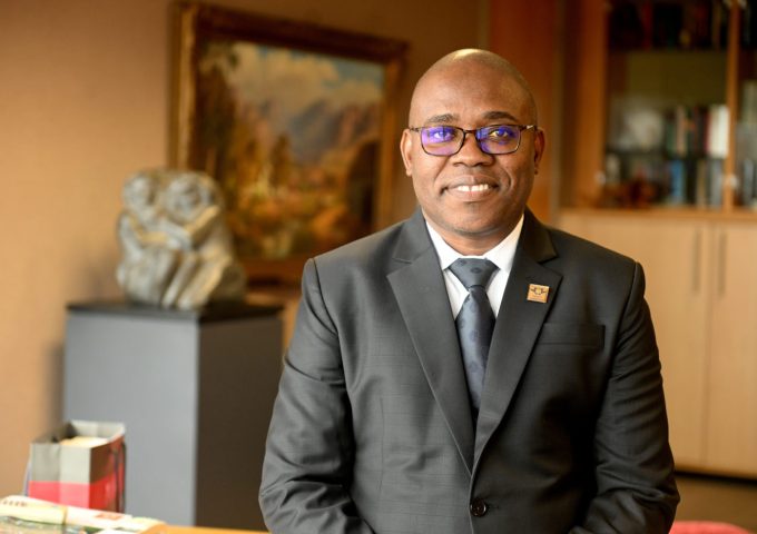 Smiling man in a dark suit and glasses seated in a warmly lit office, with bookshelves in the background.